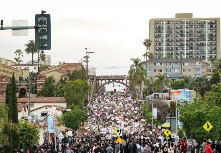 A large crowd of protesters marches down a city street holding signs. Residential buildings, palm trees, and a tall apartment complex are visible. A bridge crosses the background under an overcast sky.
