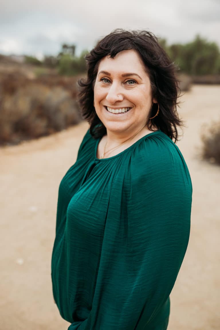A woman with wavy dark hair, wearing a green blouse, stands outdoors on a dirt path, smiling at the camera. The background is blurry with dry brush and cloudy sky.