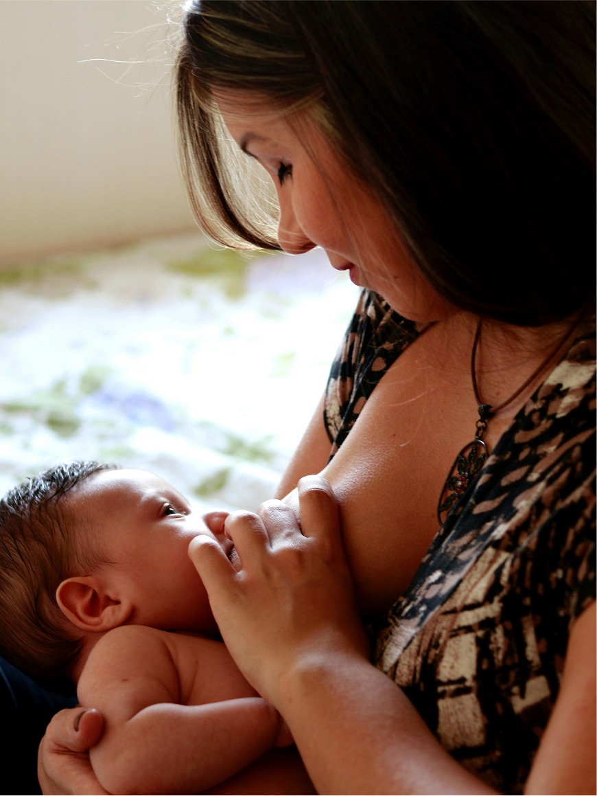 A woman sits and breastfeeds a baby, holding the child close. Both appear calm and focused on each other. The background is softly blurred, drawing attention to the mother and baby.