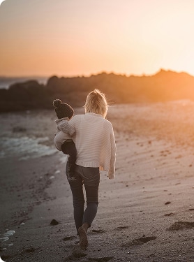 An adult wearing a white sweater and jeans walks along a sandy beach at sunset, carrying a small child in dark clothing and a beanie. Rocky formations are visible in the background.