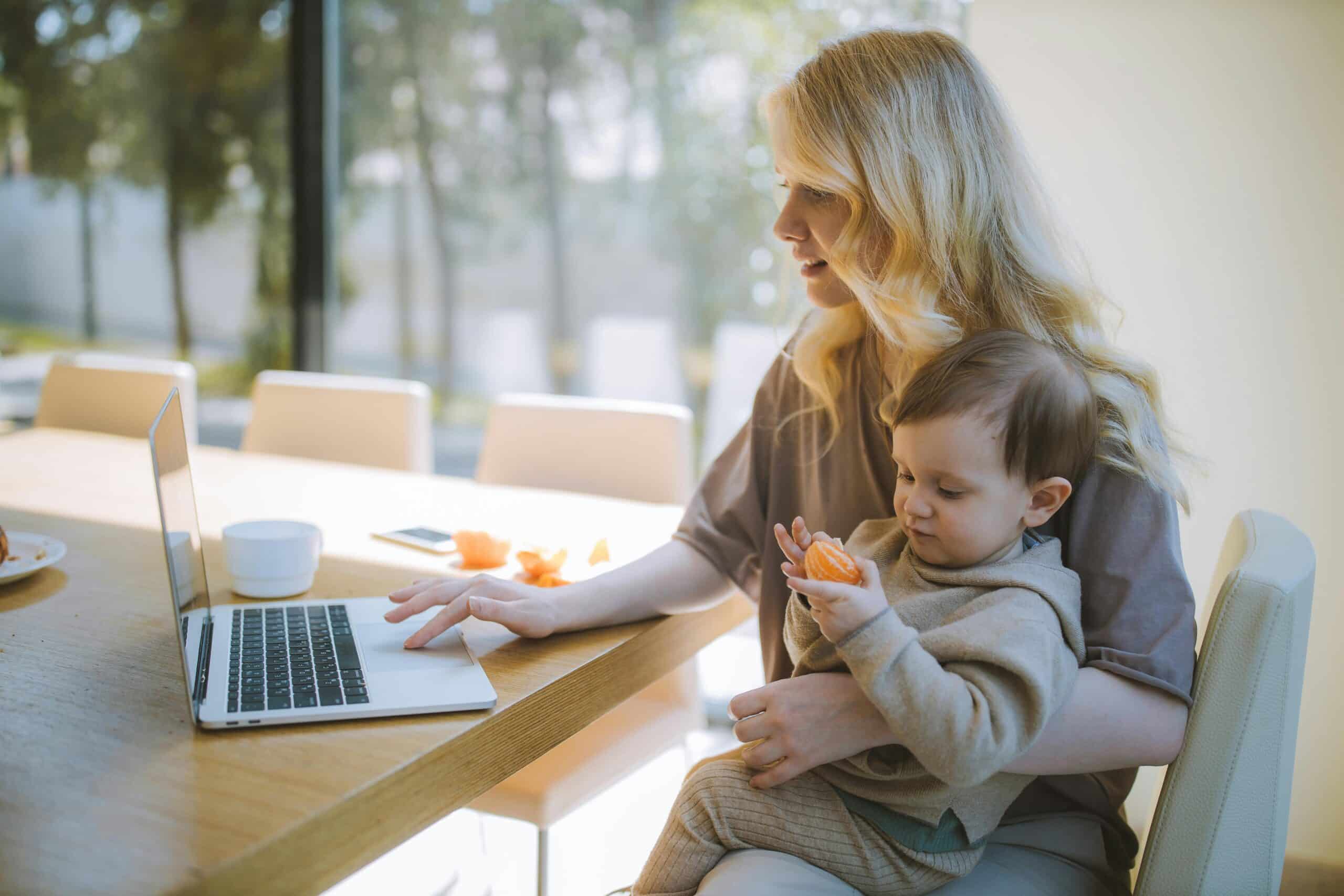 A woman sits at a table using a laptop while holding a baby on her lap. The baby is holding an orange slice. There is a cup and plate on the table, and sunlight comes through a large window in the background.