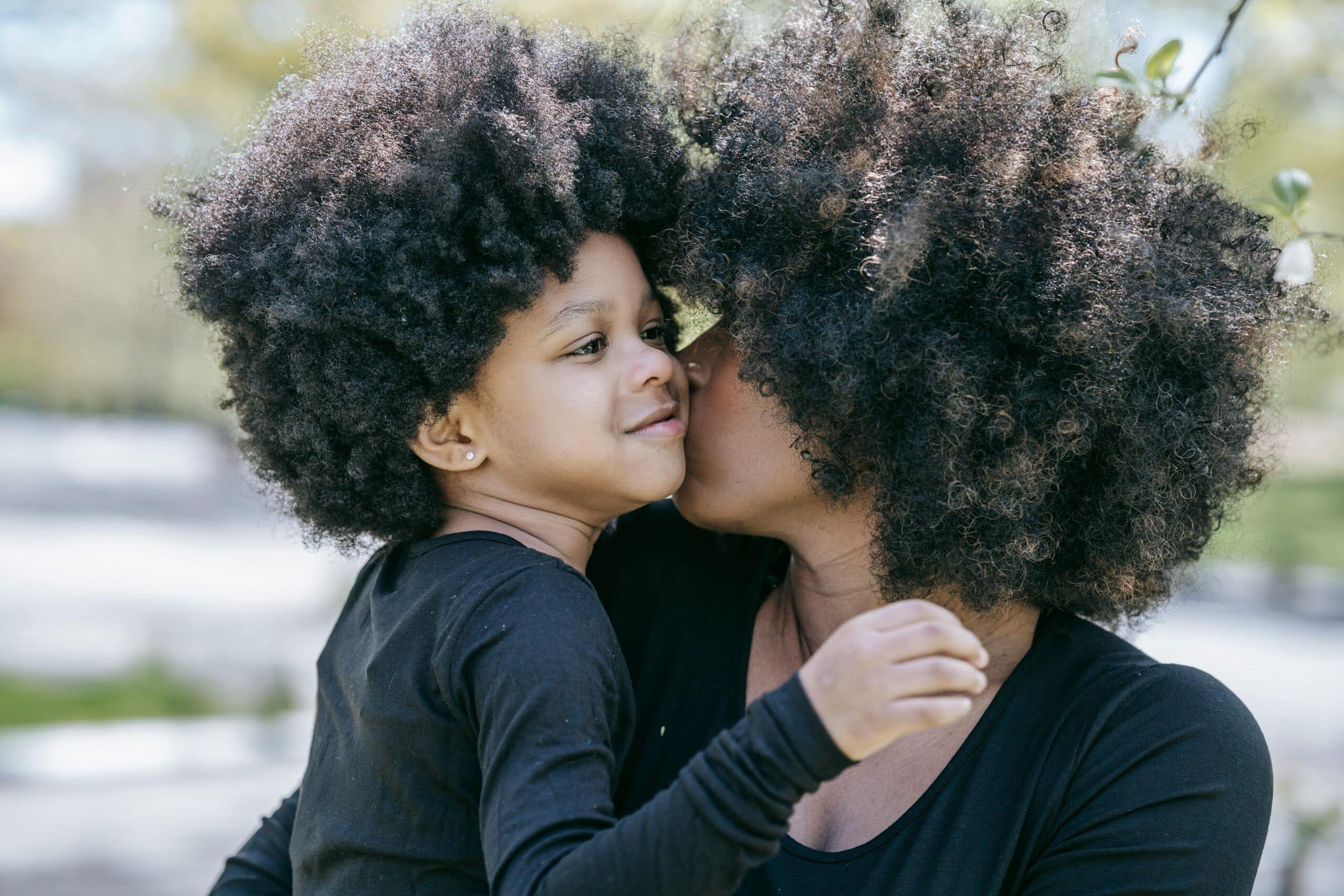 An adult holds and kisses a young child on the cheek outdoors. Both have curly hair and wear black shirts. The background is blurred, with green trees and sunlight visible.