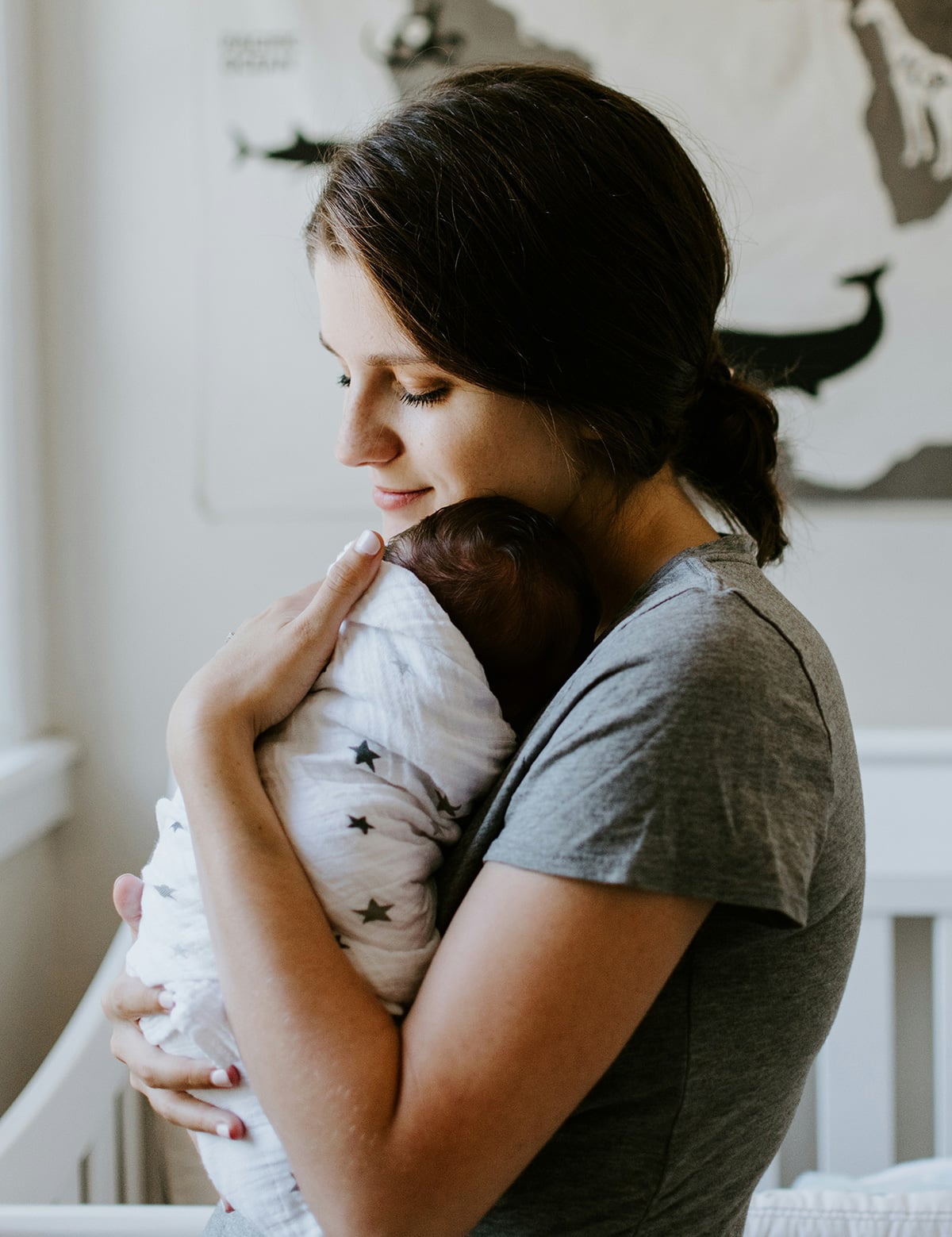 A woman in a gray shirt gently holds a swaddled baby in her arms, standing in a softly lit room with a crib and a map on the wall in the background.