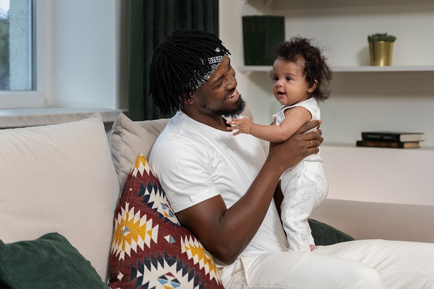 A man sitting on a couch holds a baby who is standing on his lap. Both are wearing white clothing. The man is smiling at the baby. There is a patterned pillow, books, and plants in the background.