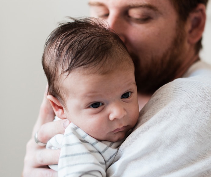 A man with a beard holds a baby against his shoulder. The baby, wearing a striped outfit, looks ahead with a calm expression. The background is neutral and out of focus.