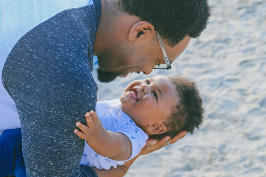 A man holds a smiling baby outdoors on a sandy surface. They are facing each other, both smiling, and sharing a joyful moment. The baby is wearing a white outfit with blue patterns, and the man is wearing glasses and a dark shirt.