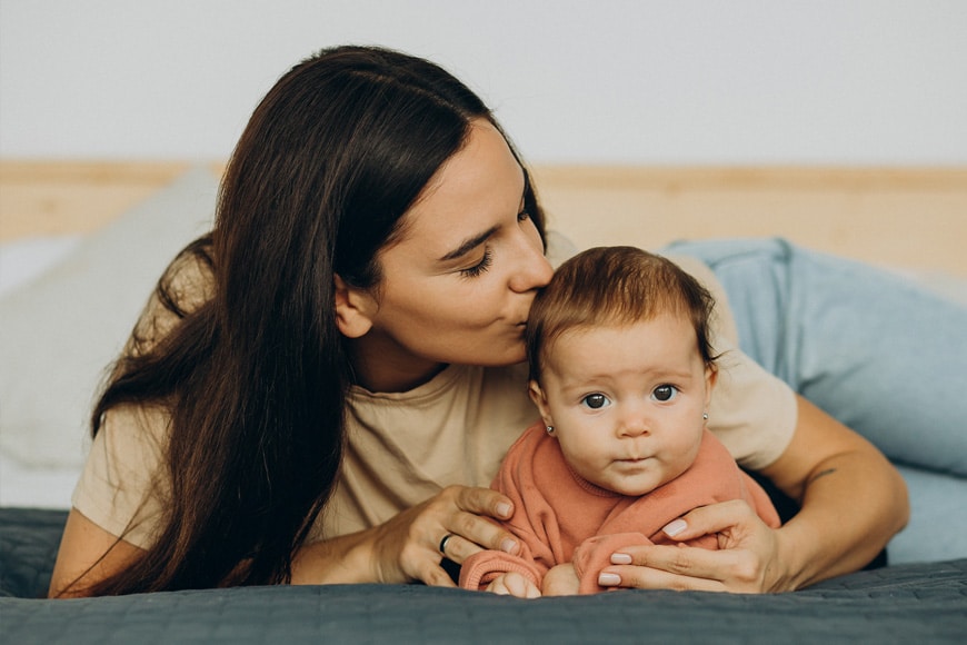 A woman with long dark hair kisses a baby with light brown hair on the head. The baby, dressed in a peach outfit, sits on a bed with gray and blue bedding, looking directly at the camera.
