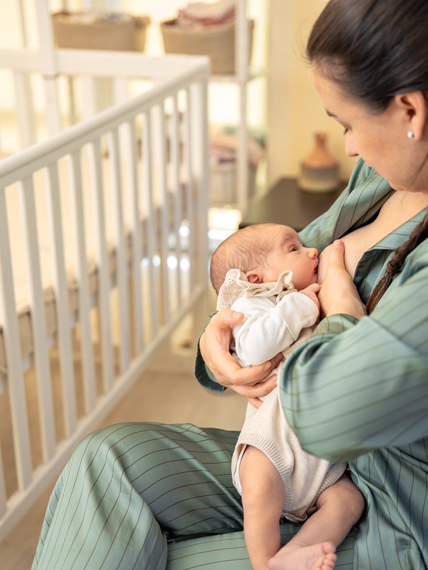 A woman in a green striped outfit is breastfeeding a baby in her arms while sitting near a white crib in a softly lit room.