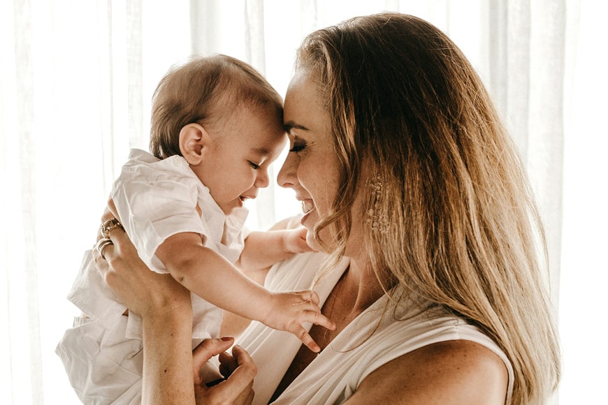 A woman holds a smiling baby close to her face. Both are dressed in white and appear happy. Light filters through a curtain in the background, creating a soft and warm atmosphere.
