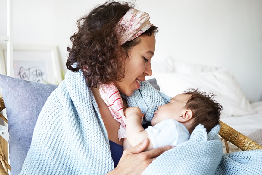 A woman with curly hair and a headscarf is breastfeeding a baby while sitting in a chair, both wrapped in a light blue knitted blanket. The background shows a white bed and framed pictures.