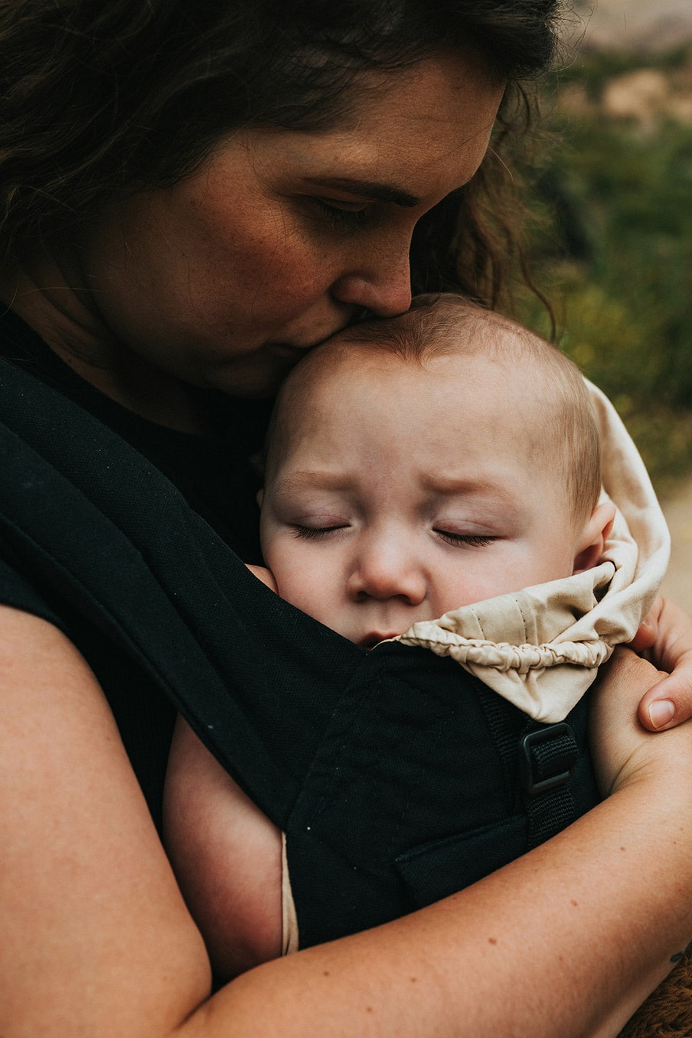 A woman holds a sleeping baby in a carrier on her chest and kisses the babys forehead. The baby has closed eyes and appears calm and relaxed. The background is blurred.