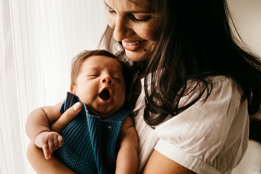 A woman wearing a white shirt holds a baby in a blue outfit. The baby is yawning, and the woman is smiling while looking down at the baby. Light comes through a window beside them.