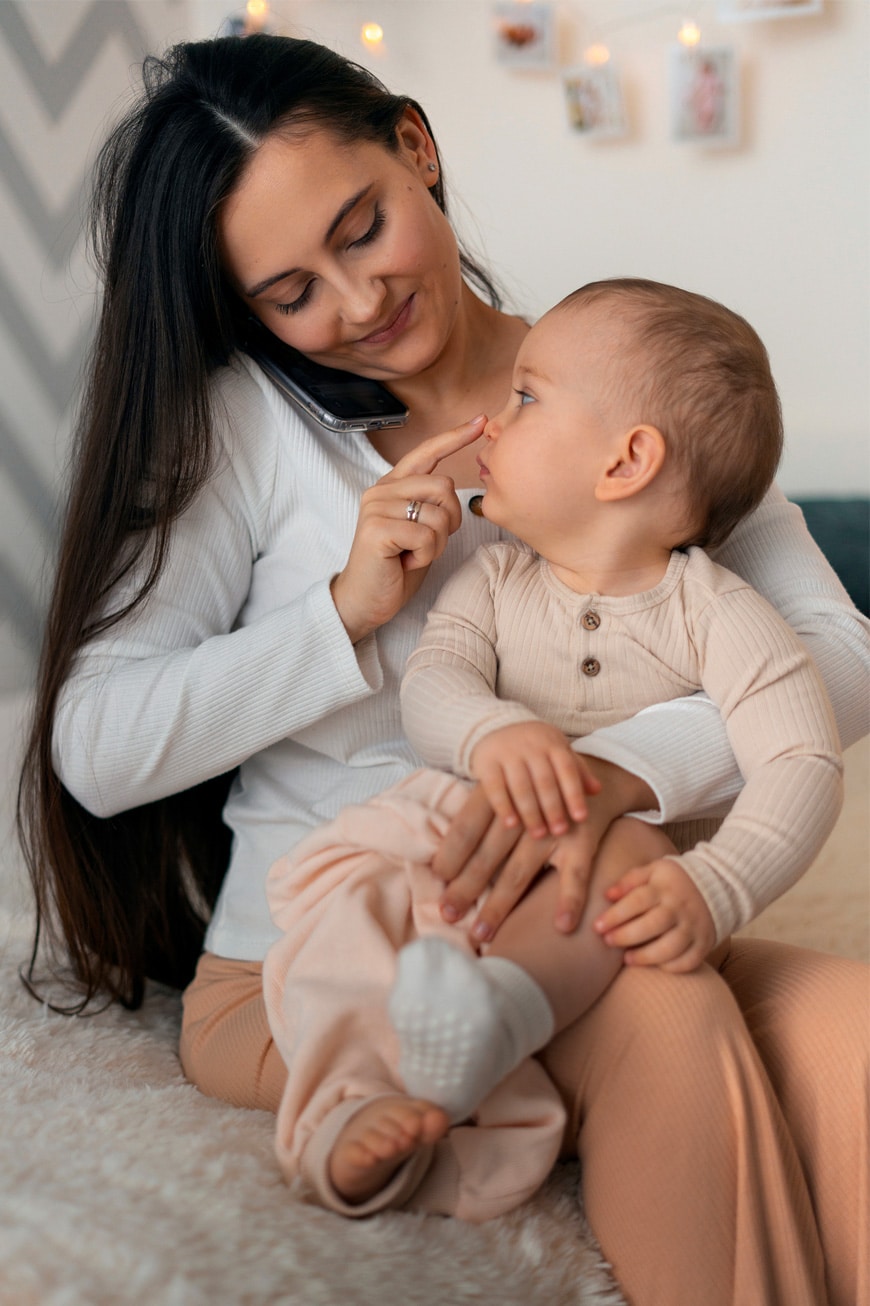 A woman sits on a bed holding a baby on her lap while talking on a mobile phone. The baby touches her face and looks at her. Both wear light-colored clothes. The background shows soft lighting and blurred photos.