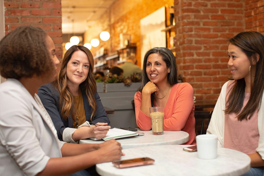 Four women sit at a round table in a cafe, talking and smiling. One woman has a notebook and pen, another has a glass of iced coffee. Exposed brick walls and warm lighting are visible in the background.